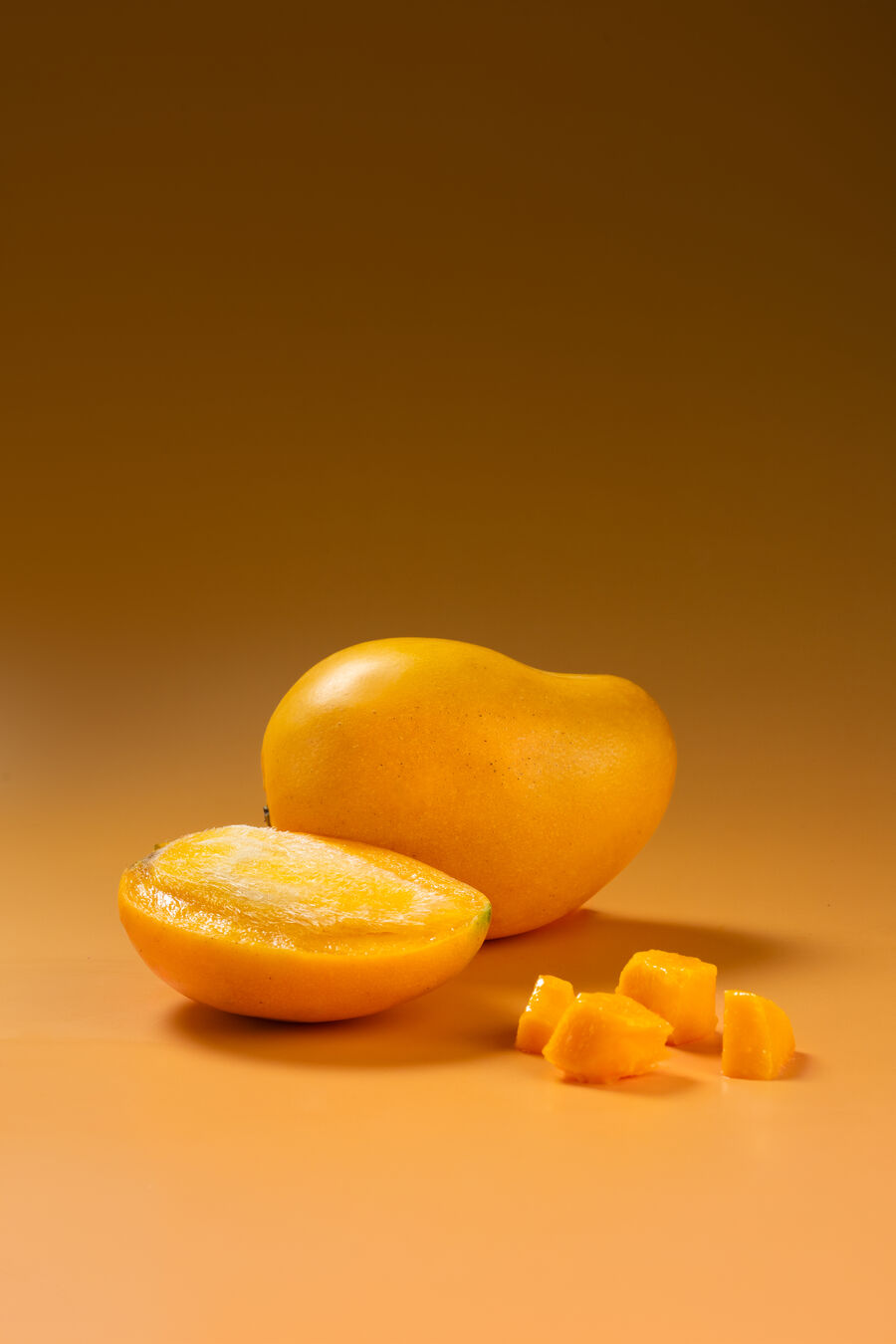 Mango Mingolo entero junto a una mitad abierta y dados de pulpa, sobre fondo ámbar de estudio.