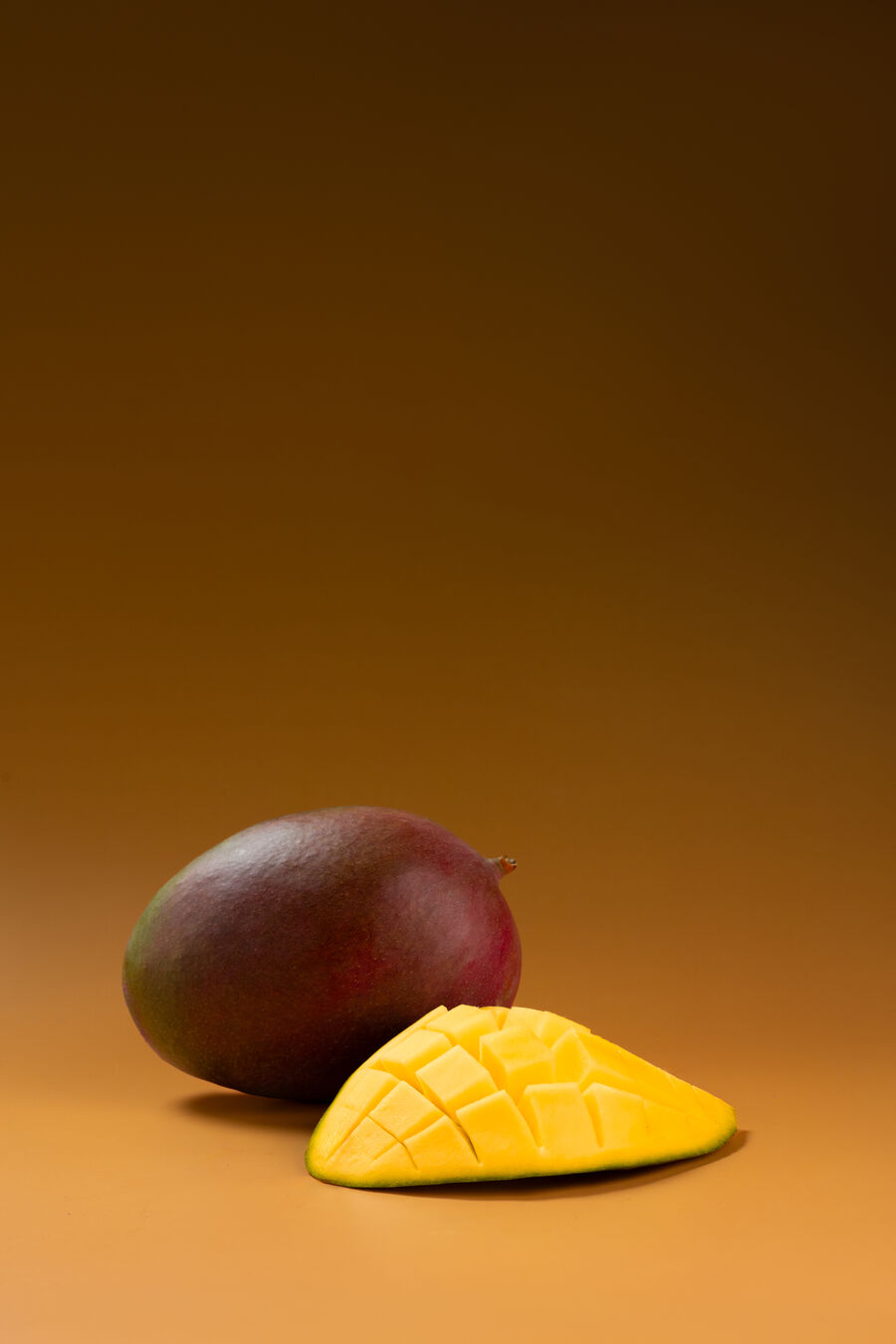 Mango Keitt entero junto a una mitad con la pulpa marcada en rombos, sobre fondo ámbar de estudio.