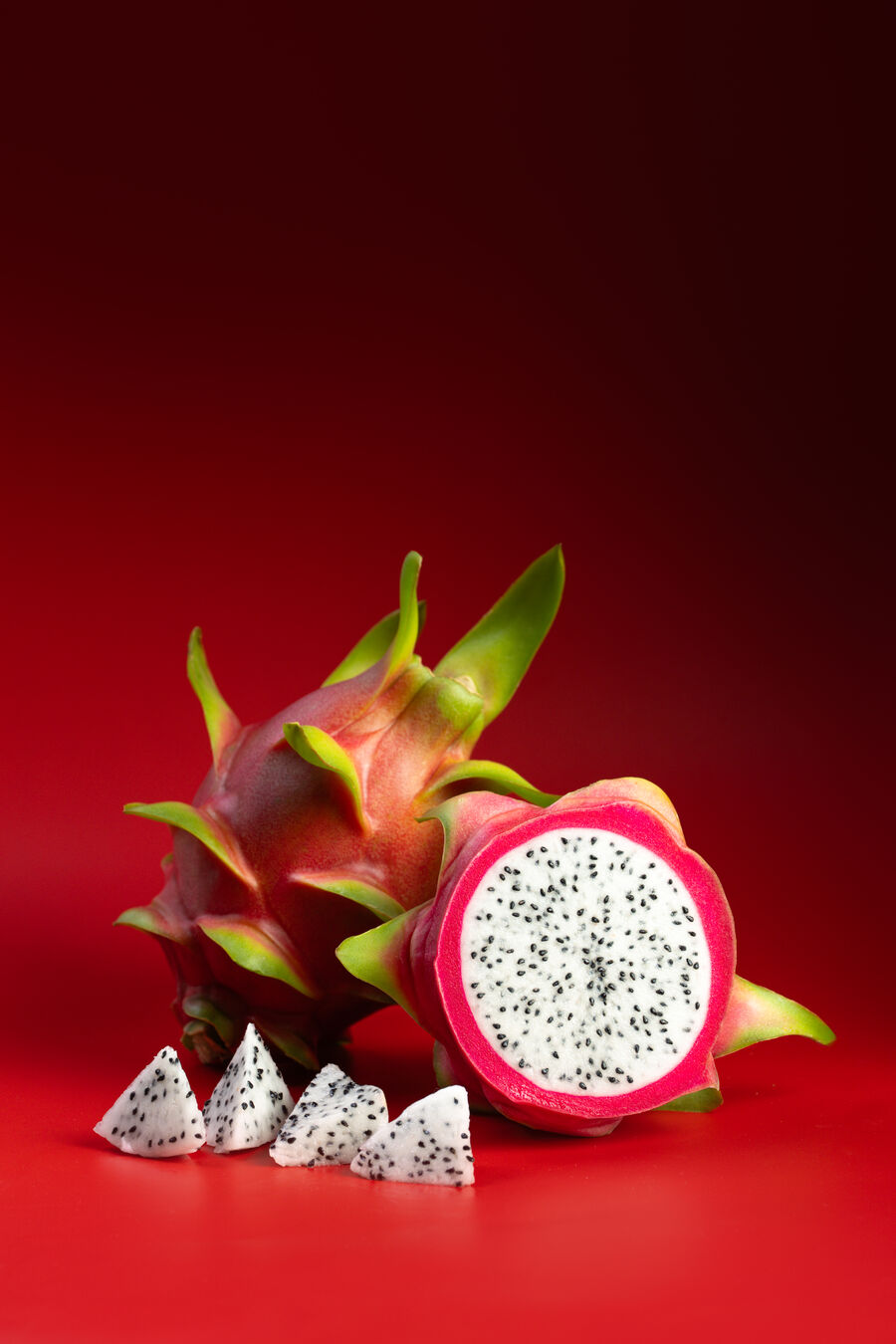 Pitahaya de piel rosada entera junto a una mitad de pulpa blanca y trozos cortados, sobre fondo rojo de estudio.