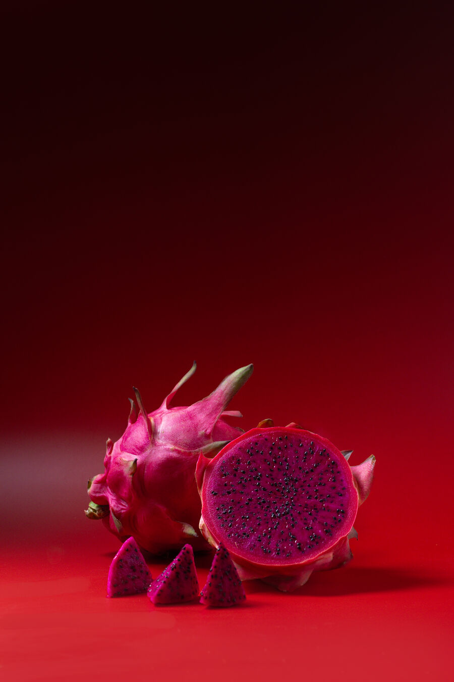 Pitahaya roja entera junto a una mitad de pulpa roja intensa y dados de pulpa, sobre fondo rojo de estudio.